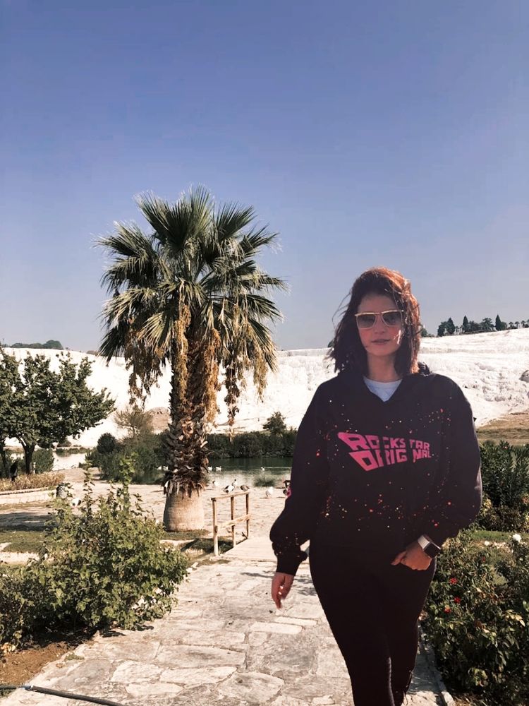 A woman wearing sunglasses and a black hoodie stands on a stone path in a park with palm trees and white rocky hills in the background.
