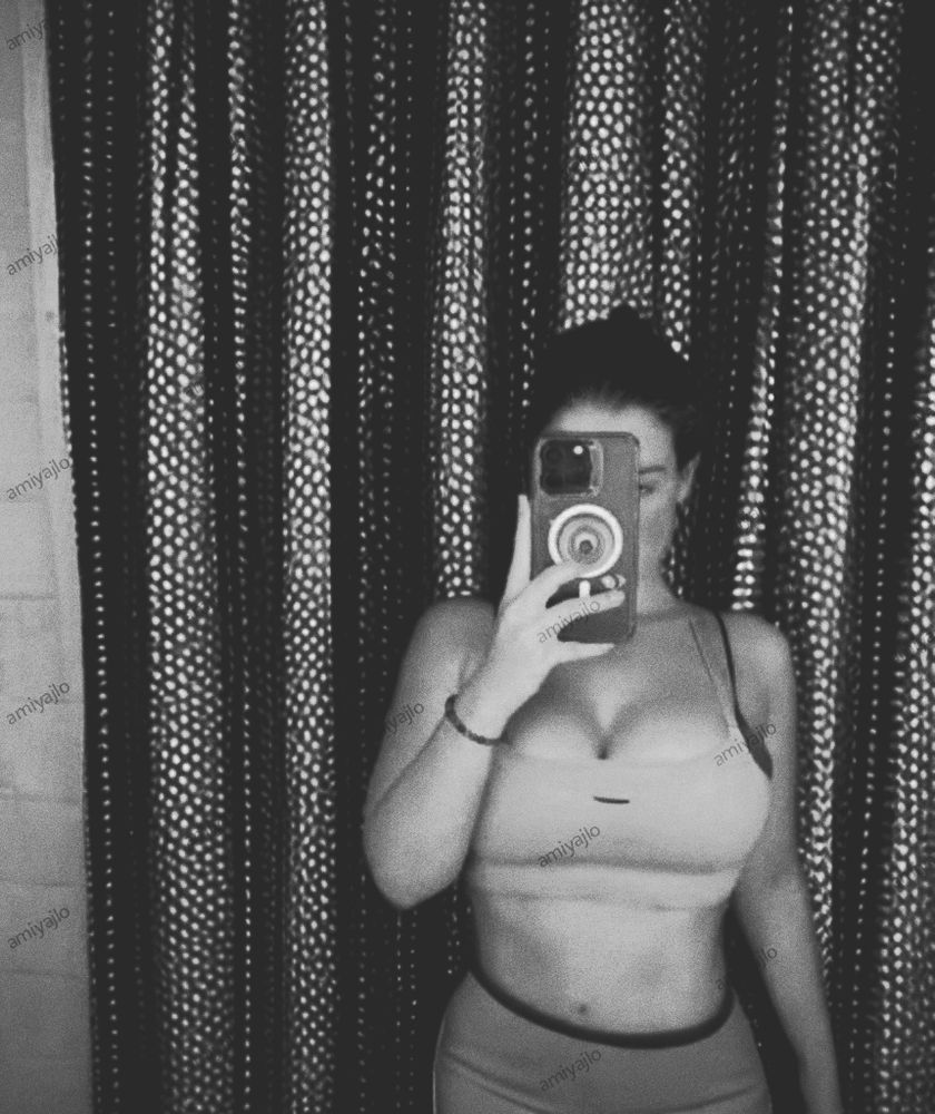 A person in a monochrome filter sportswear and shorts takes a mirror selfie, holding a phone in front of their face. Patterned curtains are visible in the background.
