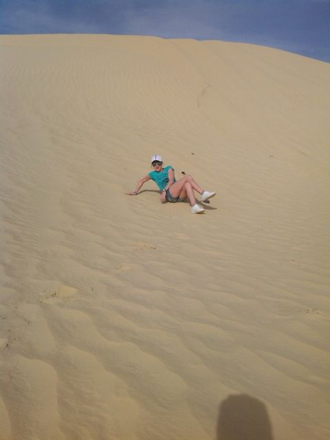 wearing a hat and shorts sits on a steep sandy dune under a blue sky, with footprints visible in the sand.