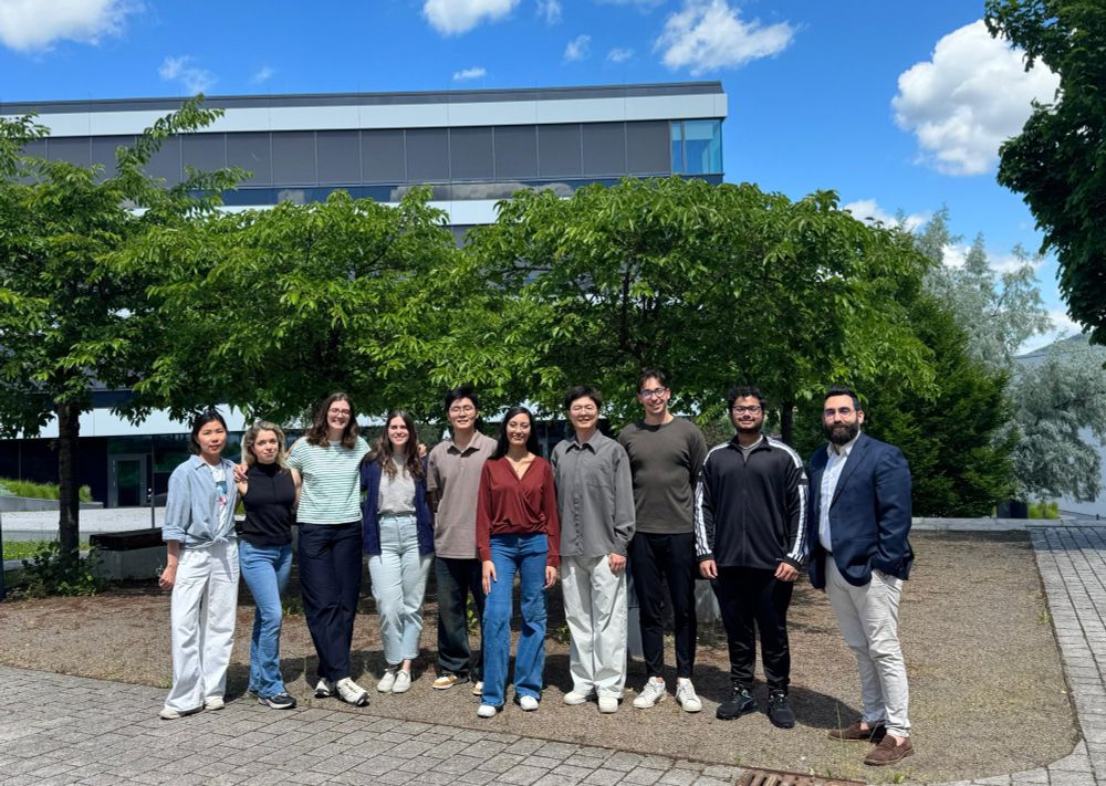 Group picture of the young international participants of the first MiCCrobioTAckle project training event on the grounds of the Leibniz-HKI in Jena. In the background are green trees and modern buildings, the sky is bright and blue with some big white clouds.