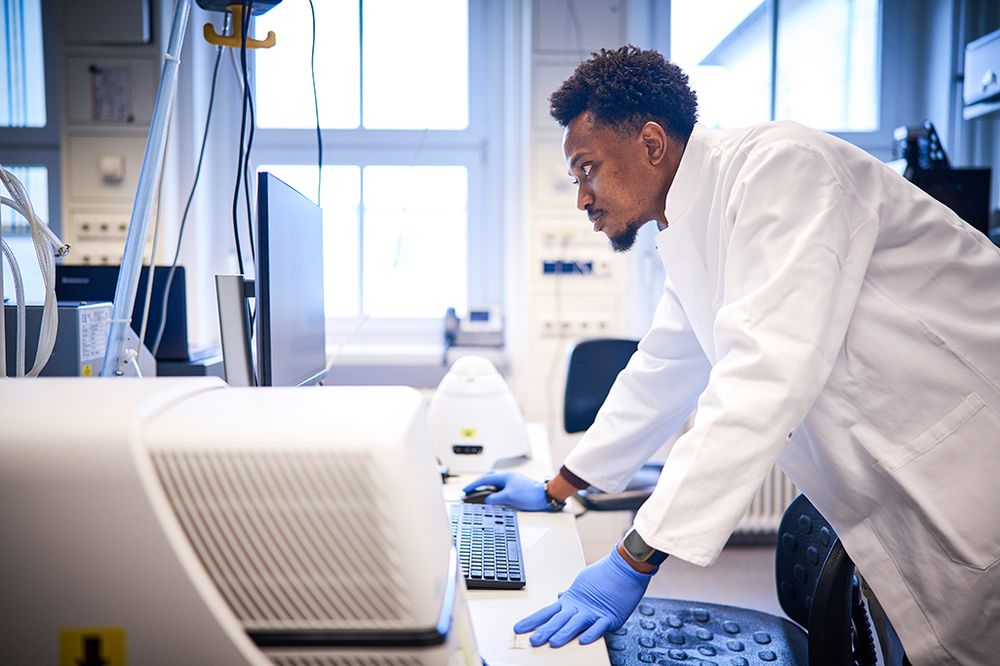 A scientist wearing a white lab coat and blue gloves leans over a desk, working on a computer in a bright, modern laboratory. Laboratory equipment and monitors surround him as natural light streams in through large windows.