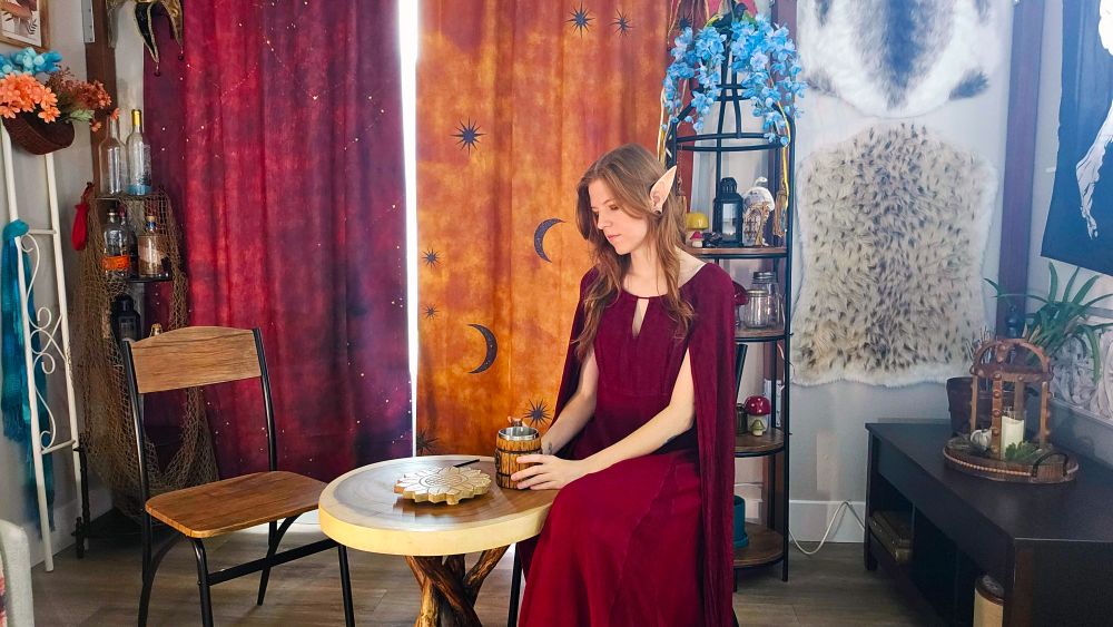 Wowzerz in a red dress. She is sitting at a table in a room decorated like a tavern.