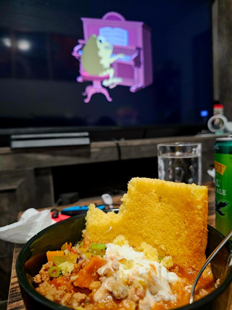 A bowl of chili and cornbread in front of a TV playing Over the Garden Wall episode 1