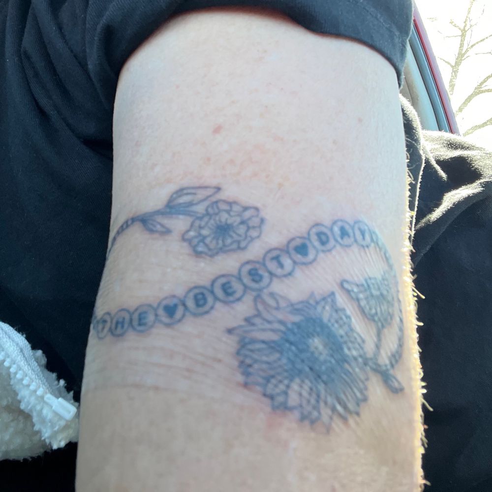 black and grey tattoo of a friendship bracelet with flowers. the friendship bracelet says “the best day”