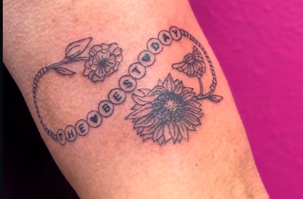 black and grey tattoo of a friendship bracelet with flowers. the friendship bracelet says “the best day”