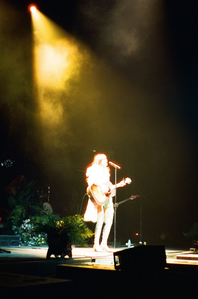 Suki Waterhouse singing and playing guitar in the spotlight.