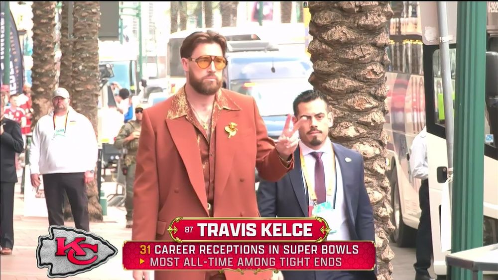 Travis Kelce wearing an orange zoot suit, a red and gold shirt with an enormous collar, yellow-brown shades, and gold flair, and flashing a peace sign