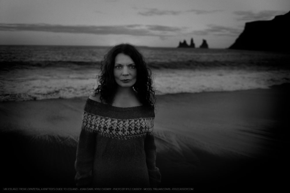 A figure wearing a cozy, off-the-shoulder sweater stands on a dark beach, with ocean waves and rocky cliffs in the background.