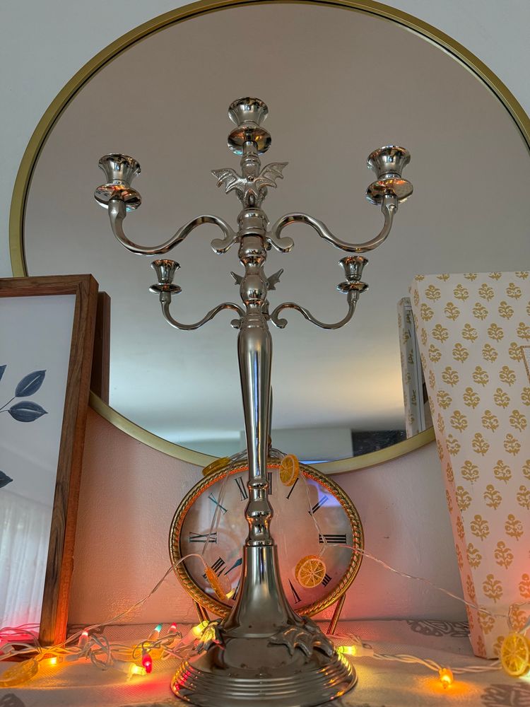 A tall silver candelabra adorned with bats sits in front of a large mirror and a clock on a mantelpiece. 
