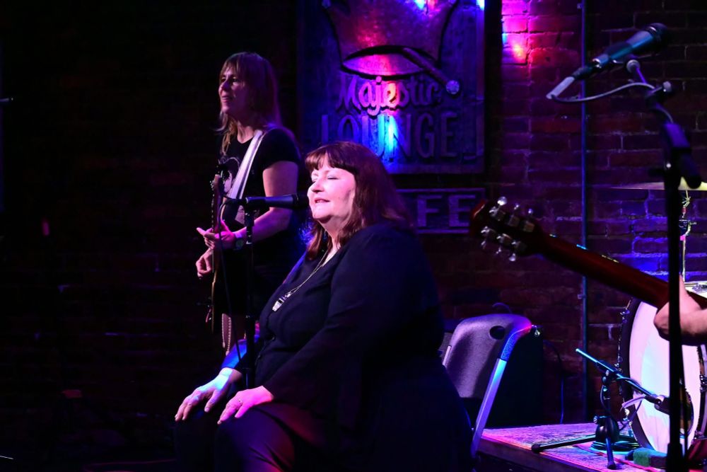 Vanessa seated center with eyes closed in front of mike, bassist Kay Stanton stands behind her. The  right edge of the photo has a guitar neck and bass drum. The stage is bathed in purple and blue light. This show was at George’s Majestic Lounge in Fayetteville Arkansas.