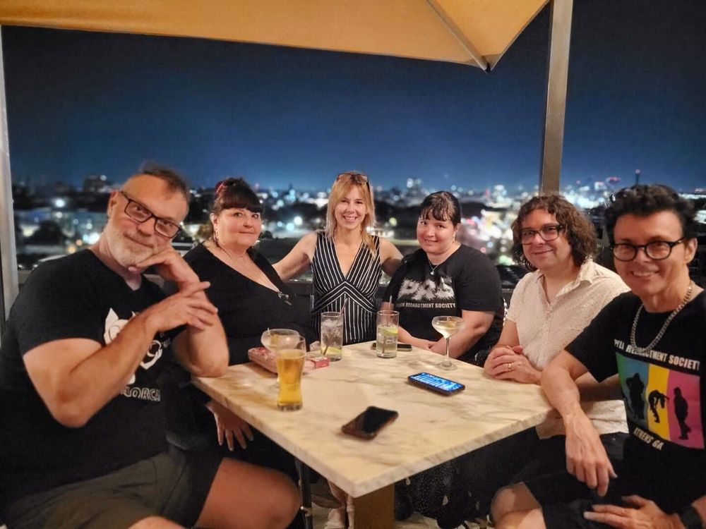 Pylon Reenactment Society, merch / roadie Hana Hay and Richard Barone seated around a table on the roof of the Thompson Hotel in Savannah, GA. The city lights of Savannah are visible behind the tired but happy group as they celebrate the end of a week long tour which covered 6 Southeastern US cities in 7 days and logged 2500 miles.