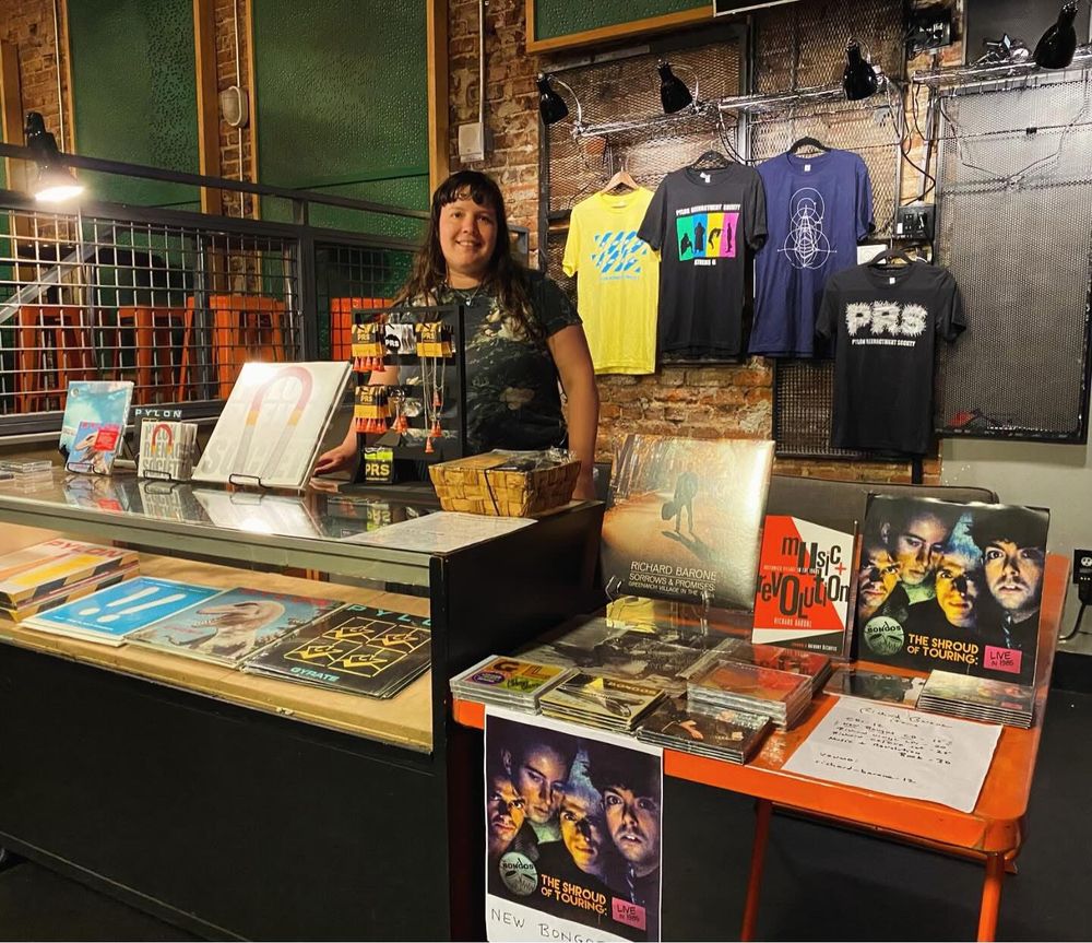Hana Hay behind the merch booth at the Saturn Birmingham. Left glass case Pylon and Pylon Reenactment Society recordings and merch including earrings, on the right is an orange table with Richard Barone and The Bongos recordings, merch and one of Richard’s books. Back wall with PRS and Pylon T shirts - pRS yellow with blue safety stripes, PRS silhouettes black T shirt with green, blue, yellow and magenta design, Navy blue Pylon T shirt with white design of a Pylon, Black T shirt with Silver magnetic design logo. 