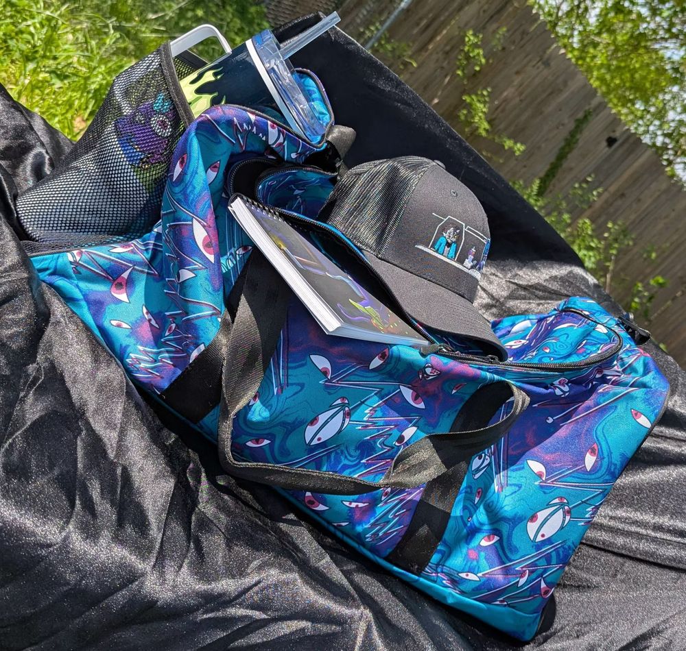 A photograph of several pieces of merch from my store.  A duffle bag patterned with eyes and lighting that comes in two colour ways, a silly trucker hat with truckers on it, a notebook with one of my illustrations on the cover, and a big travel mug.