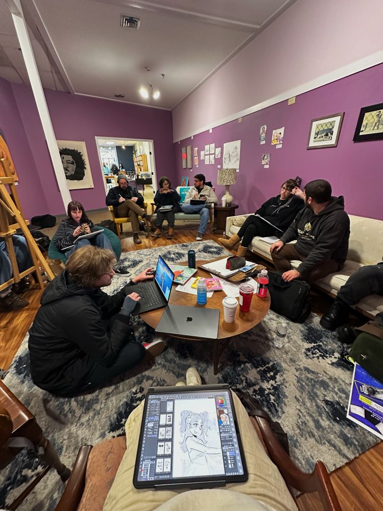 Several artists gathered around a table in the purple room at The Cordial Eye, working on their comics. Sketchbooks, laptops, and art supplies fill the room. Some artists are deep in concentration, while others chat and share ideas. The room has a cozy, creative energy.