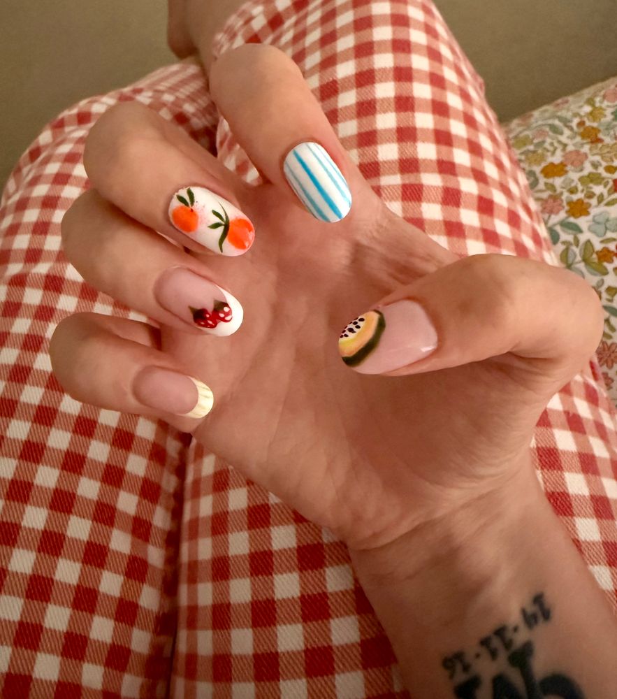 A set of nails on one hand, each one different. The thumb is plain with part of the inside of a passion fruit on the corner, the index finger has blue and white stripes, the middle finger has a white background with oranges on, the ring finger is plain white a white tip and strawberries on and the little finger is plain with a yellow and white tip 