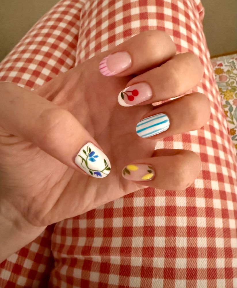 A set of nails on one hand, each one different. The thumb has a white background with blue flowers on a green stem, the index finger is plain with a pink striped tip, the middle finger is plain with a white tip and some cherries, the ring finger has blue and white stripes and the little finger has lemons on 