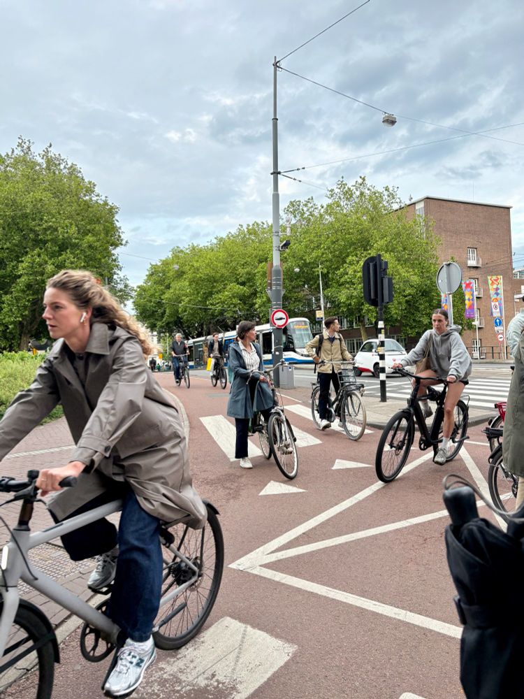 A busy crossroad with many people riding bicycles from different directions, some trees, and a tram in the background under a cloudy sky.