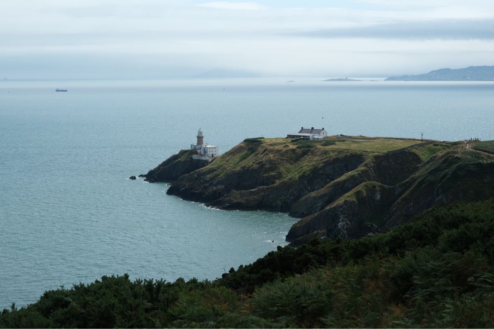 A white lighthouse with a black roof perched on a distant cliff overlooking the sea. The sea is a deep blue, with some clouds in the sky.
