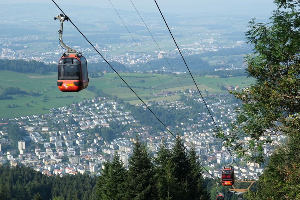 A mountain cable car system with two red cars, one higher than the other, against a panoramic view. The background features a sprawling cityscape with numerous buildings amidst patches of greenery and trees. In the distance, the horizon is visible where the sky meets the land.