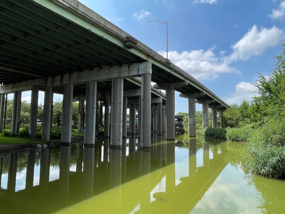 A freeway overpass crossing above a large body of standing water (Flushing Creek). The water is very green and marshy. This is in the northeast part of Flushing Meadows.