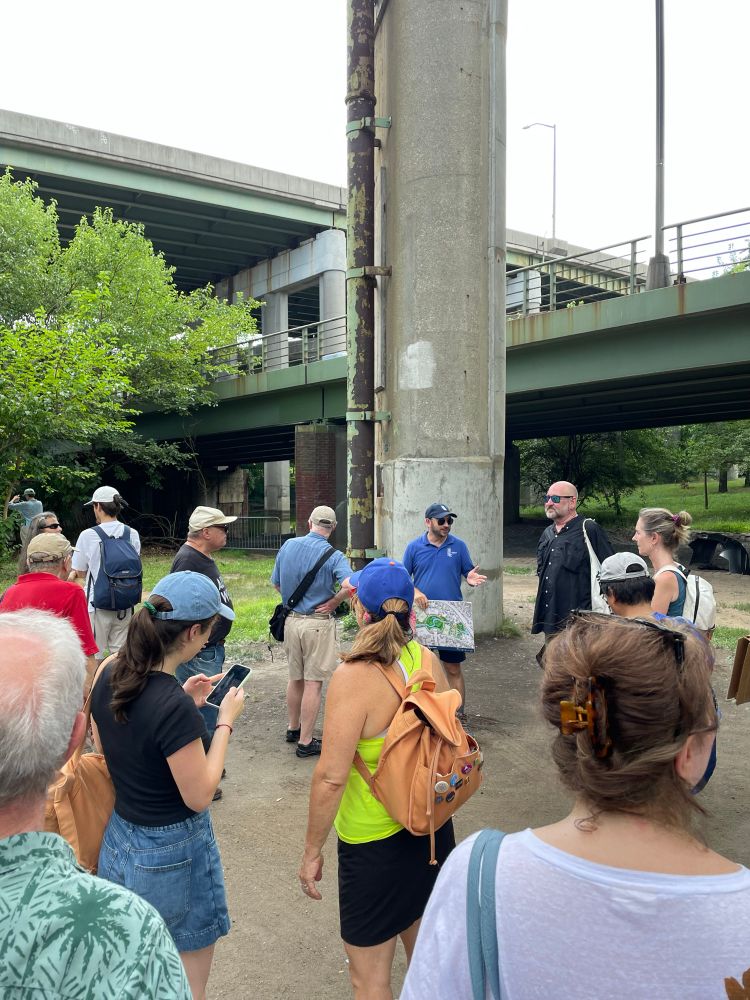 Walking tour group standing in the park. A freeway overpass crosses overhead and a large metal pipe (scupper) that drains runoff from the freeway can be seen on the supporting pillar. The end of the scupper can’t be seen but when it rains, it just discharges on the ground where the group is standing.