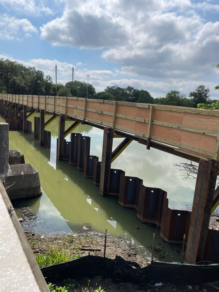 A walkway, under construction, crossing over a very green and marshy body of water. Beneath the bridge are metal tide gates preventing water flow. This is Tide Gate Bridge in Flushing Meadows.