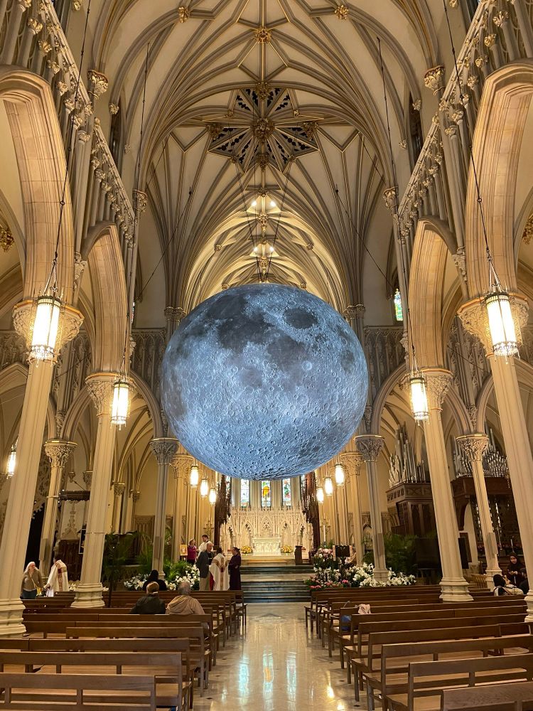 Interior of a cathedral church in the French Gothic style from the center aisle facing the altar. In the crossing is suspended an enormous sculpture of the moon. About 20 feet in diameter, the moon fills most of the volume in the high pointed arch of the center of the building.