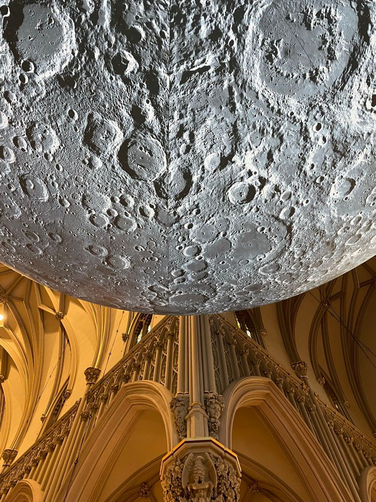 An oblique view of the moon sculpture from underneath and corner detailing of the crossing in the background. In the Gothic style, the arches in the stonework are pointed at the top and the columns are carved so as to look like bundles of thinner columns. At the bottom of the photo is one of the capitals with carvings depicting an eagle surrounded by foliage.