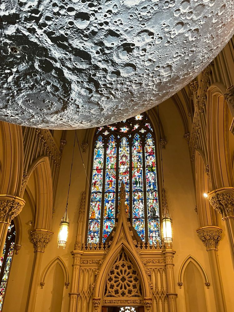 One of the large stained glass windows at the end of the transept with a wishbone shaped arch in stone in front of it. The moon sculpture appears floating in the upper left corner of the image in the foreground.