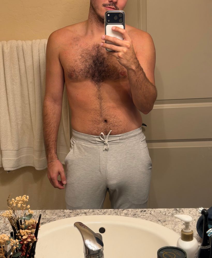 A bathroom mirror self of louie in grey sweat with thick vpl. He is tan, with dark, curl chest hair.