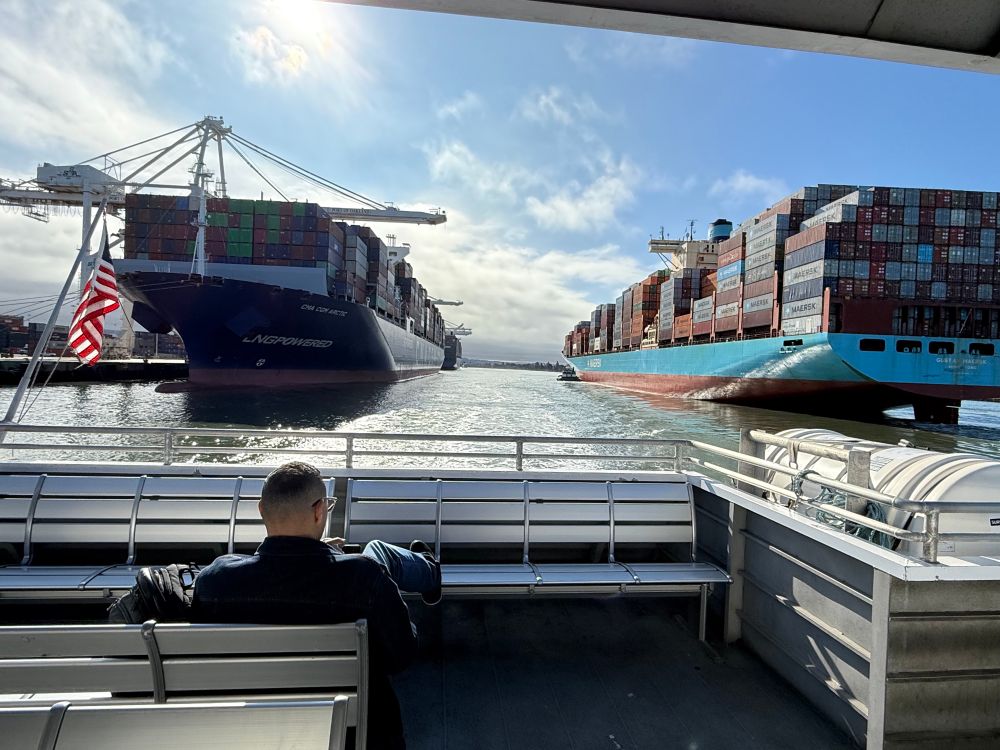 View from the back of a ferry with an American flag passing between two large container ships. 