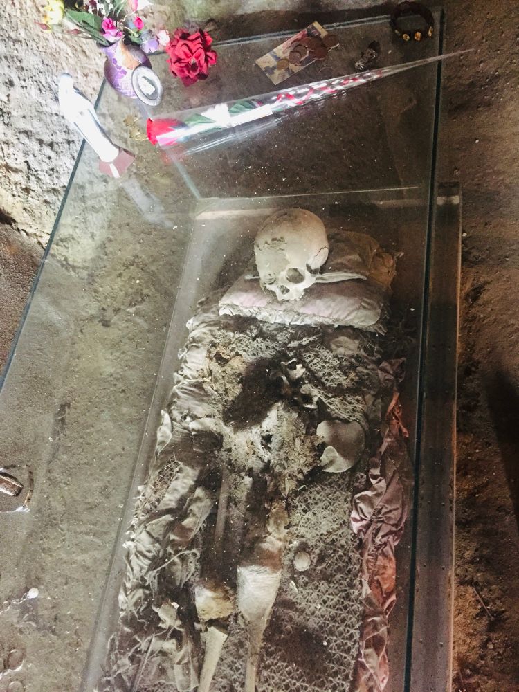 A glass coffin in the catacombs of Cimitero delle Fontanelle in Naples, with a human skull on a worn pillow and other human bones on a faded blanket; flowers, coins, jewelry, and statues of saints on the coffin.
