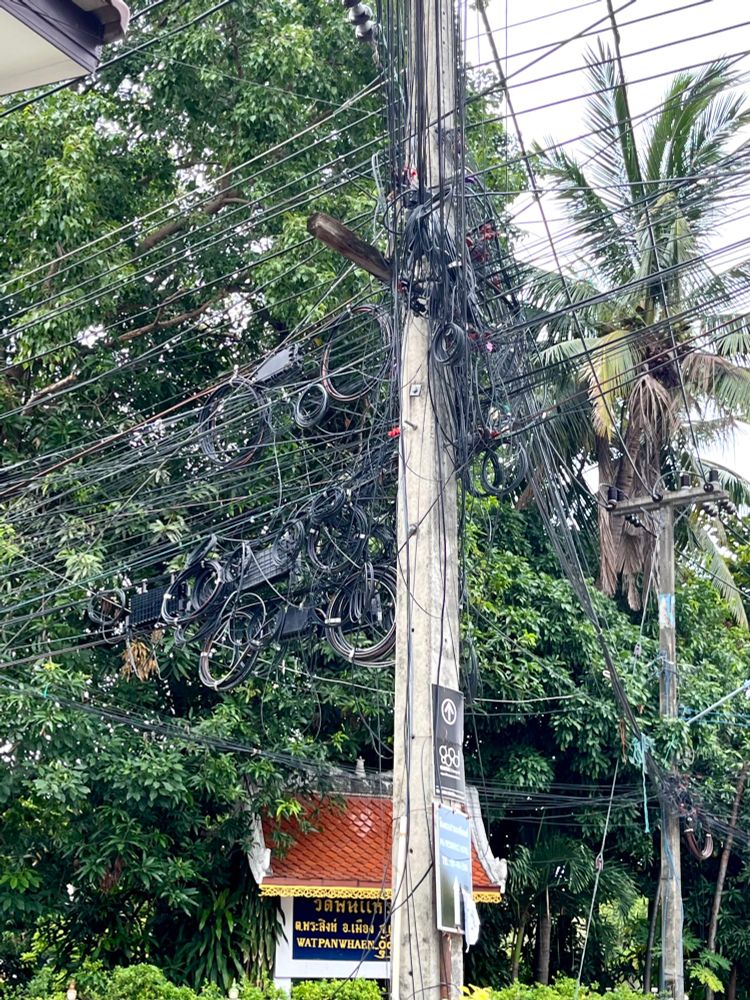 Ein Strommast mit Dutzenden Stromkabeln, die in alle möglichen Richtungen abgehen; im Hintergrund das Eingangsschild eines thailändischen Tempels vor großen Bäumen.
