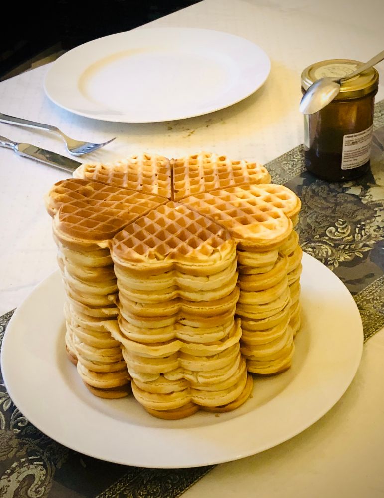 Im Vordergrund ein weißer Teller mit einem Stapel frisch gebackener Waffeln, im Hintergrund ein weiterer Teller mit Besteck, sowie ein Glas Karamell mit Löffel darauf. 