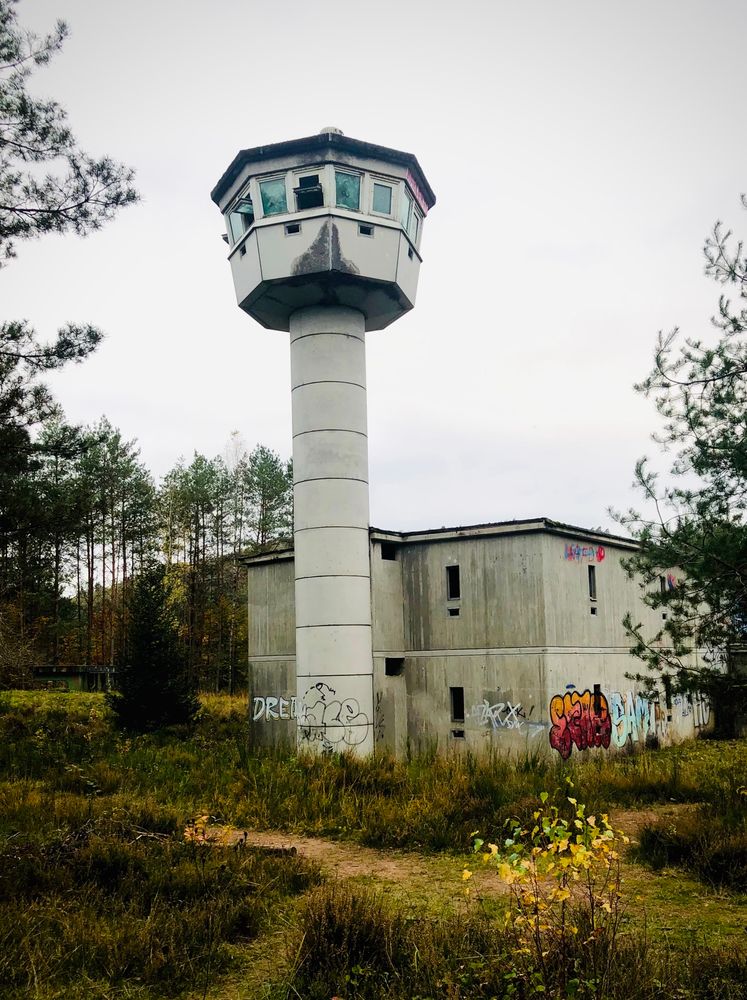 Ein verlassenes Militärgebäude mit Überwachungsturm, an den Wänden mit Graffiti besprüht, umgeben von Wald.