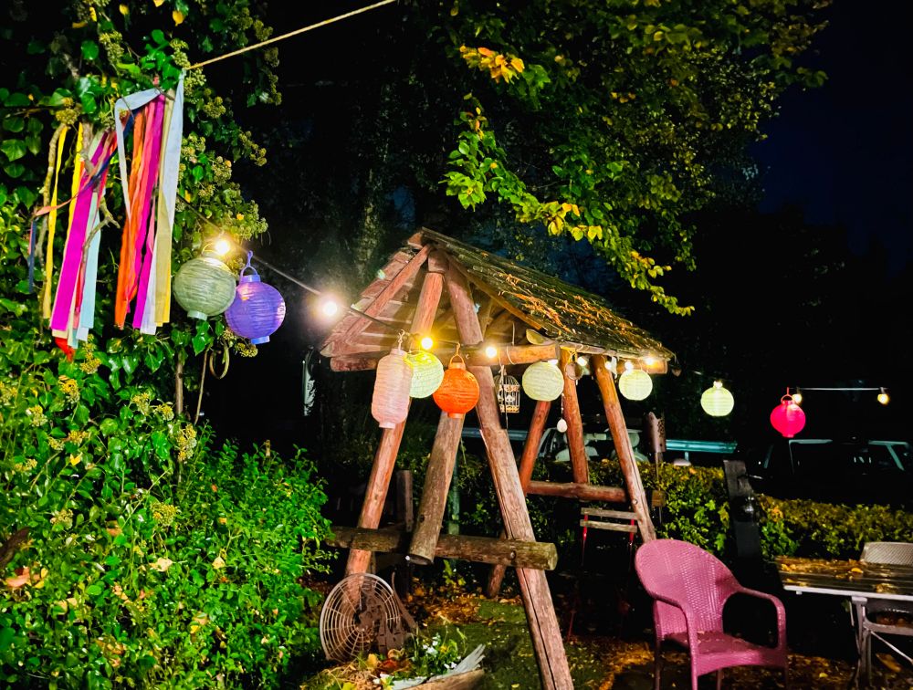 Blick auf einen Biergarten bei Nacht mit bunten Laternen, Papierstreifen und witterungsbedingt leeren Stühlen und Tischen.