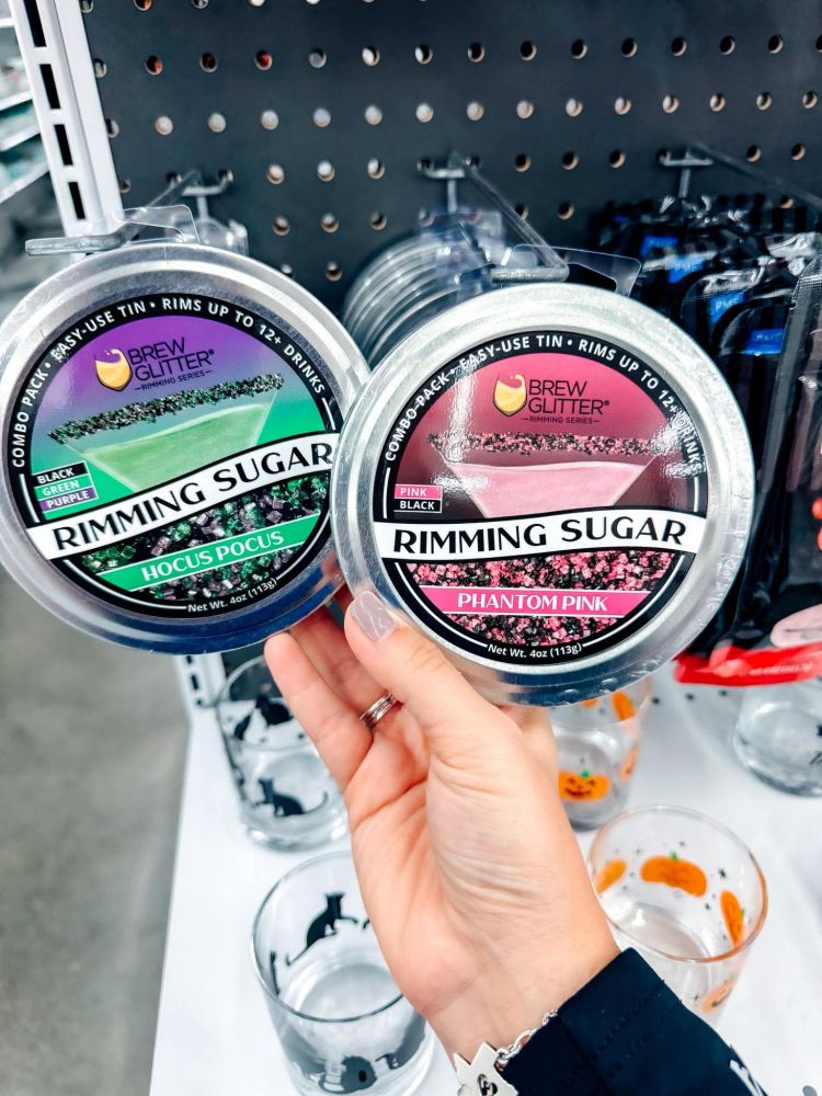 A close-up shot of a person's hand holding two plastic containers of cocktail rimming sugar. The one on the left is labeled "Hocus Pocus" and contains a mix of green, black, and purple glitter sugar. The one on the right is labeled "Phantom Pink" and contains a mix of pink and black glitter sugar. In the background, there are several glasses decorated with Halloween themes like pumpkins and black cats.
