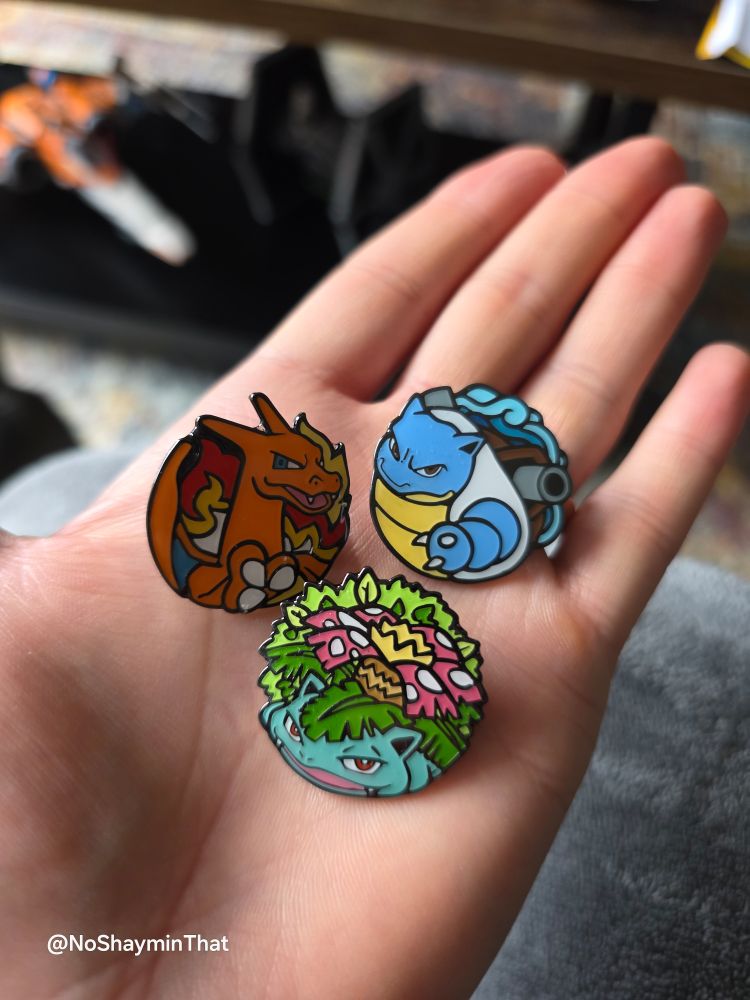 My open palm holds three colorful enamel pins. The pins are of the final evolved forms of the Kanto starter Pokémon: Charizard, a fiery orange dragon; Blastoise, a blue turtle with cannons on its back; and Venusaur, a large green plant-dinosaur hybrid. The pins are arranged in a triangle formation, with Charizard and Blastoise at the top and Venusaur at the bottom. The background is out of focus, showing a glimpse of what appears to be a sofa or blanket. In the bottom left corner of the image, the text @NoShayminThat is visible, which appears to be a social media handle.
