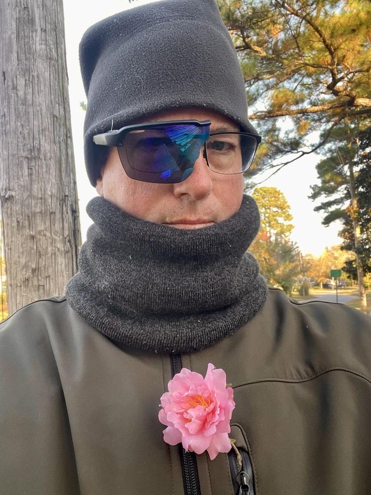 Selfie picture of me on the morning walk. I’m wearing a sage green zip up jacket. Around my neck is a gray tube scarf bunched up to just below my mouth. On my head is a gray watch cap. In the front chest pocket is a pale pink camellia flower. I’m wearing regular glasses, but on the right side is also half a broken pair of sunglasses that has a large dark blue tinted lens that I found curbside.