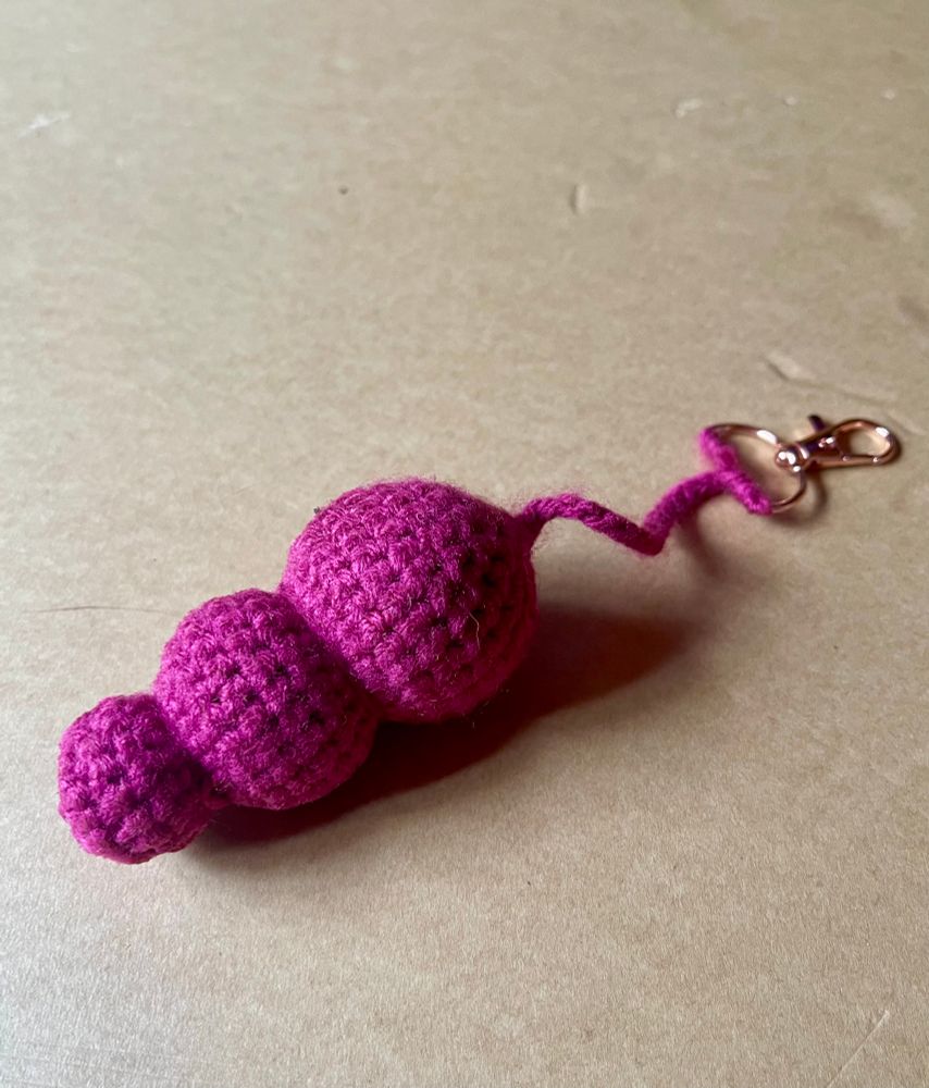A pink crochet amigurumi keychain shaped like an anal sex toy with three balls that increase in size. The keychain is attached to a rose gold clasp. It is on a neutral beige background. 