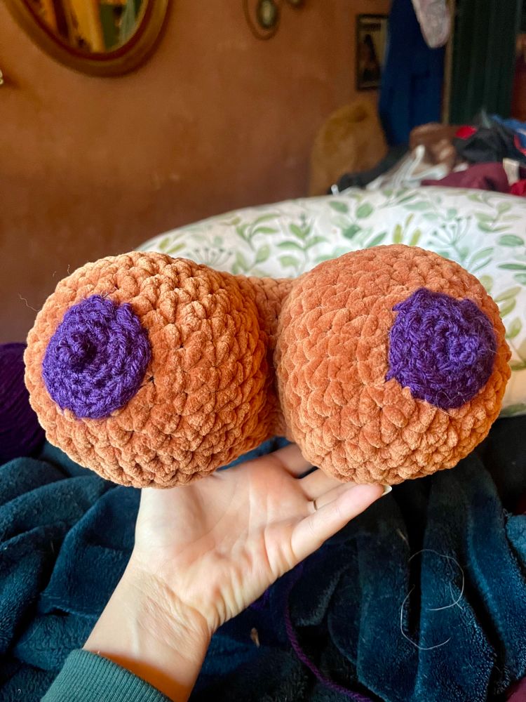 A persons hand holding an orange crochet amigurumi heart, facing the camera from a top down angle the heart looks like a pair of breasts with two crochet purple nipples 