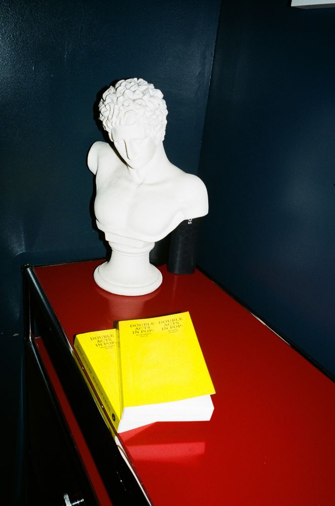 Two copies of "Double Acts in Pop" on a red table next to a classical sculpture of a bust.