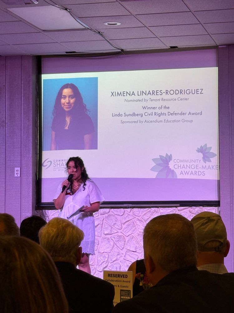 Ximena speaking to a room full of people. The Screen behind her says "Ximena Linares-Rodriguez Nominated by Tenant Resource Center. Winner of the Linda Sundberg Civil Rights Defender Award." 