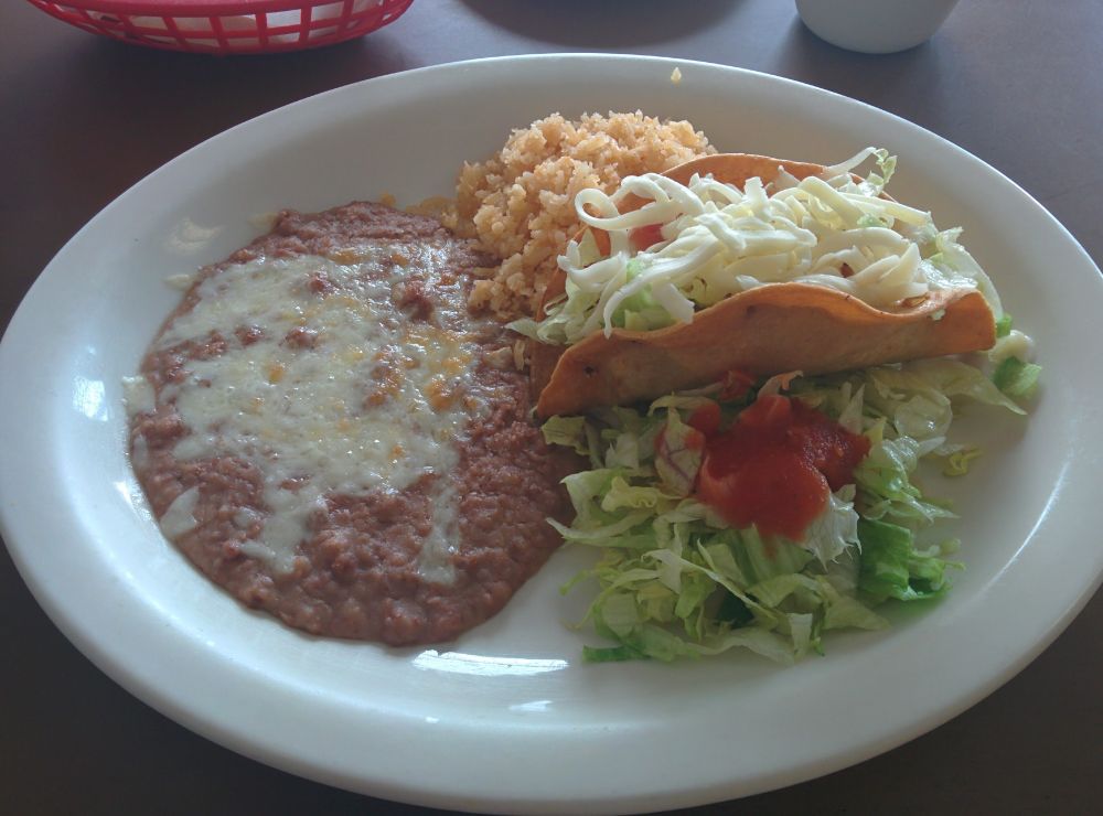 Plate with hard-shell taco, Mexican rice, refried beans with cheese, and a red basket in the background.