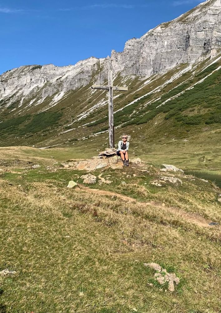 Eine Person sitzt unterhalb  eines Gipfelkreuzes, im Hintergrund Gebirge 