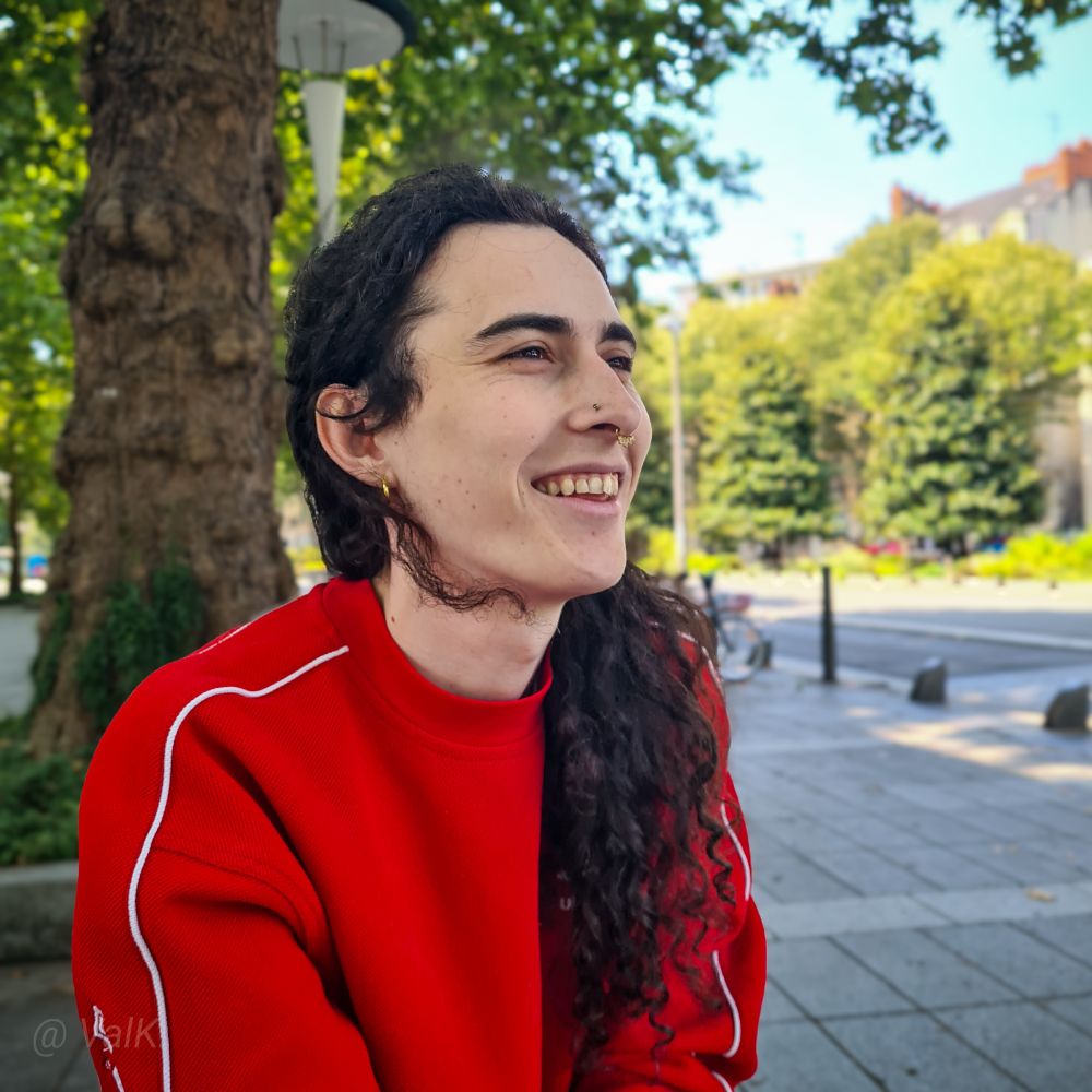 Photo de Pia avec un grand sourire, sous un arbre, en pull rouge, ses longs cheveux noirs bouclés qui tombent en cascade sur ses épaules 