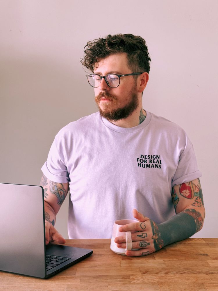 A man sat at a table, using a laptop, wearing a t-shirt that says 'Design for Real Humans'