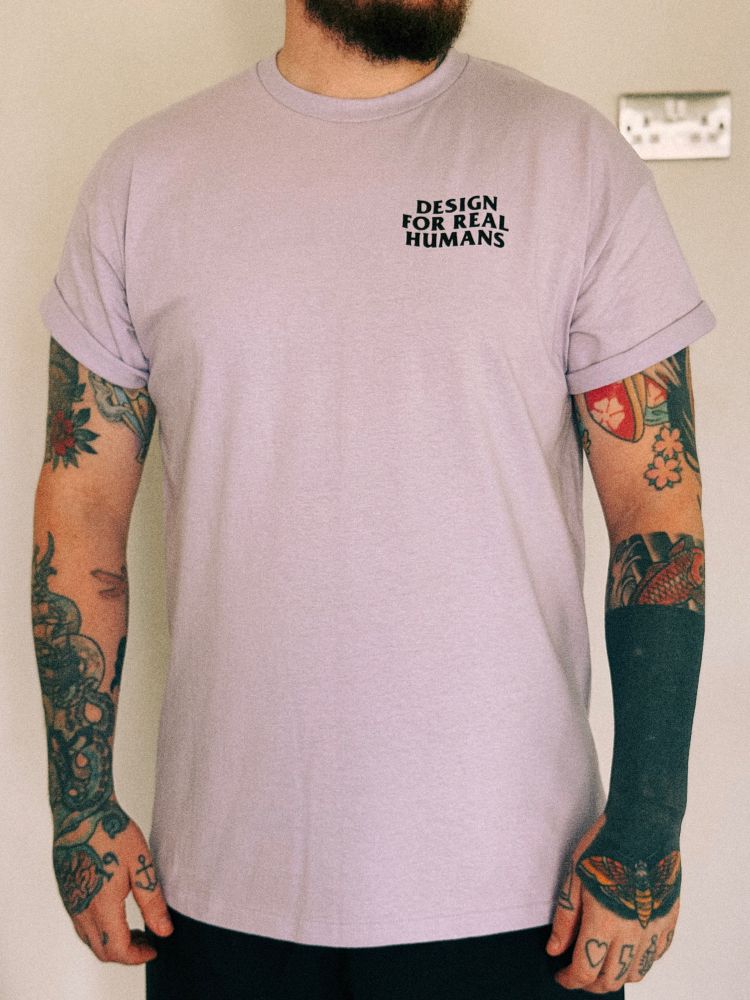 A tattooed man wearing a lavender t-shirt with ‘design for real humans’ in a wavy font on the left chest of the t-shirt