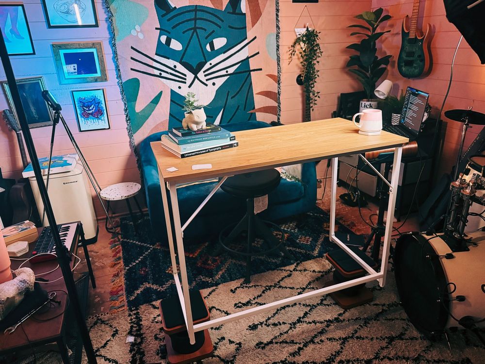 behind the scenes shot of a video recording setup. a desk is balanced precariously on speaker stands. the room is messy but has a very nice cat tapestry hanging on the wall.