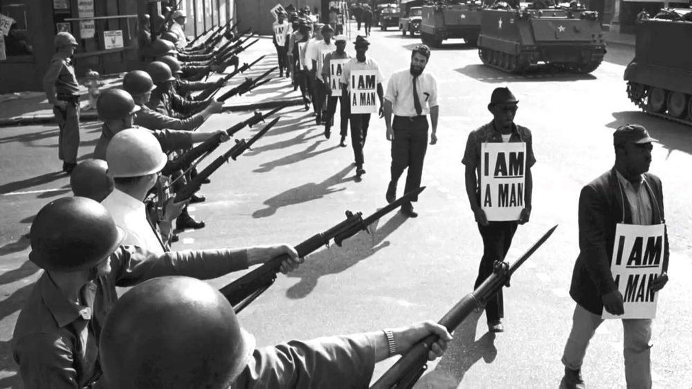 Black men wearing boards that say "I AM A MAN" walking past US soldiers pointing bayonettes at them.